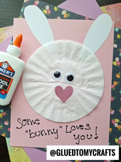 A handmade bunny craft made from a white coffee filter, pink paper, googly eyes, a pink heart nose, and white paper ears—perfect for coffee filter crafts for kids. Elmer’s glue and decorative paper are nearby. Text reads: “Some ‘bunny’ loves you!”.
