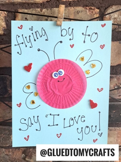Handmade cupcake liner card crafts on blue paper with “flying by to say ‘I love you!’” written in black. A pink cupcake liner decorated as a bug with googly eyes, wings, and heart stickers. @GLUEDTOMYCRAFTS is at the bottom.