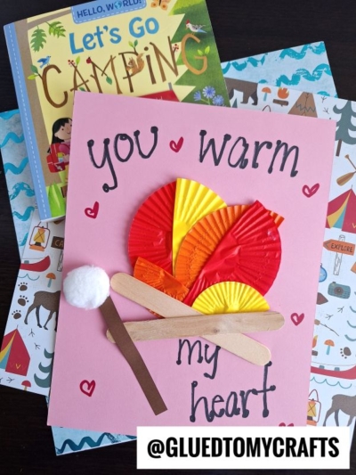 A handmade cupcake liner card crafts project with "You warm my heart" text, featuring a campfire made from paper, cupcake liners, craft sticks, and a cotton ball marshmallow on camping-themed books and scrapbook paper.