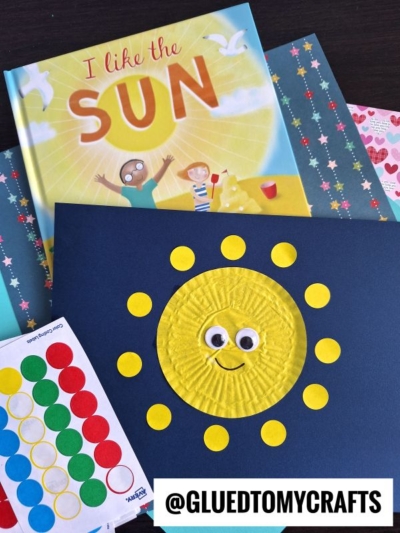A children’s book titled I Like the Sun, a sheet of star-patterned paper, colorful dot stickers, and a sun craft made from a yellow cupcake liner with googly eyes—perfect for cupcake liner card crafts on blue paper.