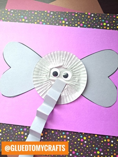 A handmade elephant cupcake liner card craft featuring googly eyes, a folded paper trunk, and gray paper ears, displayed on a pink and star-patterned background.