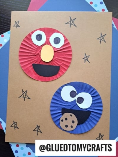 Two cupcake liners, one red and one blue, are transformed in this cupcake liner card crafts project into Elmo and Cookie Monster with paper cut-out faces and a cookie, displayed on brown paper decorated with hand-drawn stars.