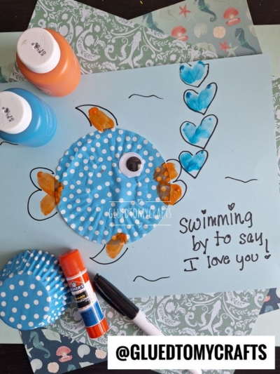 A handmade fish craft on blue paper uses a blue polka-dot cupcake liner for the fish’s body, orange-painted fins, a googly eye, and heart bubbles. Perfect for cupcake liner card crafts, this project says “Swimming by to say I love you!”.