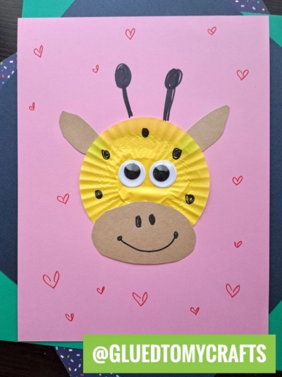 A cute giraffe face made from a yellow cupcake liner, paper cutouts, and googly eyes is glued to pink paper with red hearts—an adorable example of cupcake liner card crafts. The username @GLUEDTOMYCRAFTS is displayed at the bottom.