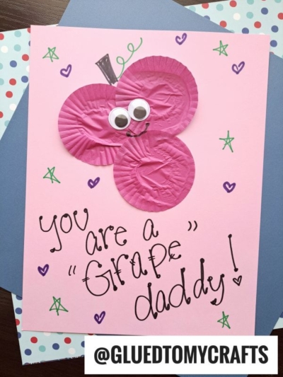 A pink cupcake liner card crafts project featuring two purple liners shaped like grapes, googly eyes, a drawn stem and leaf, plus a handwritten "You are a Grape Daddy!" message with stars and @GLUEDTOMYCRAFTS at the bottom.
