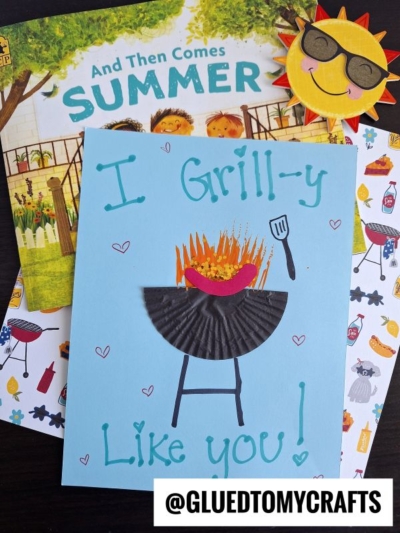 A handmade cupcake liner card crafts a paper grill and food, reading I Grill-y Like You! sitting on a summer-themed book and stickers. A smiling sun craft is nearby, with the handle @GLUEDTOMYCRAFTS visible.