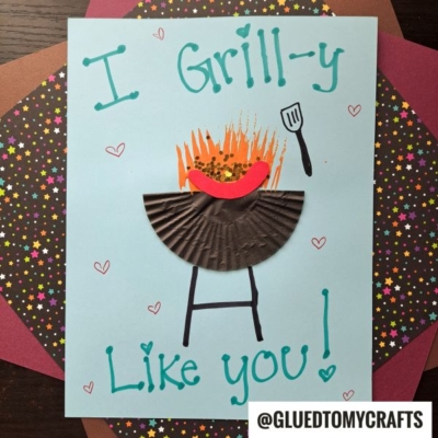 A handmade HANDPRINT FATHER'S DAY card featuring a grill made from a brown cupcake liner, orange flames, and a red paper hot dog. It says I Grill-y Like You! on a blue background with colorful star paper behind.