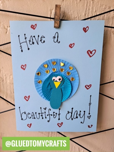 A handmade cupcake liner card features a blue peacock with gold accents and red hearts. The card reads "Have a beautiful day!" and is clipped to a string. Perfect for cupcake liner card crafts lovers! @GLUEDTOMYCRAFTS is labeled at the bottom.