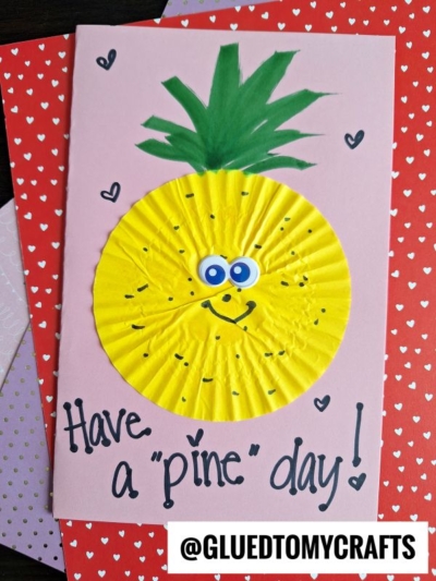 A handmade cupcake liner card features a yellow liner decorated as a smiling pineapple with googly eyes and a green paper crown. The card says, Have a pine day! and sits on a red heart-patterned background—perfect for fun card crafts.