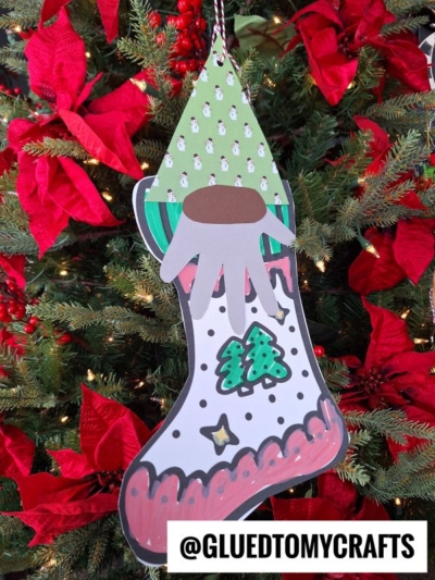 A handmade gnome stocking ornament decorated with green, red, black, and white patterns hangs on a Christmas tree with red poinsettias. The bottom right corner has the text @GLUEDTOMYCRAFTS.