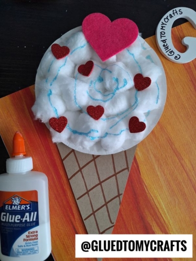 A charming cotton ball ice cream cone craft with glittery red heart decorations and a felt heart on top. Elmer’s glue and a GluedToMyCrafts tag sit nearby. @GLUETOMYCRAFTS is at the bottom for easy inspiration.