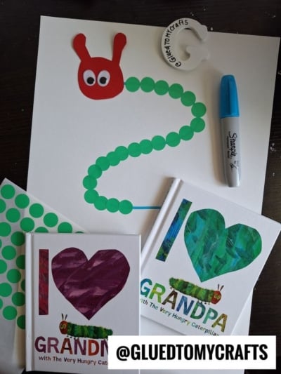 A HUNGRY CATERPILLAR CRAFT made with green dot stickers and a red paper head sits beside a Sharpie marker, glue bottle cap, and two The Very Hungry Caterpillar themed books. @GLUEDTOMYCRAFTS is written at the bottom.