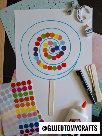 A spiral lollipop art project made with colorful dot stickers on white paper, surrounded by glue, craft sticks, a black marker, star stickers, coding label crafts supplies, and colorful papers. @GLUEDTOMYCRAFTS text at the bottom.