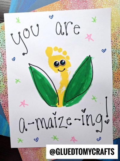 A handmade card features a yellow footprint styled as corn with green husks, stars, and hearts—a cute handprint Father's Day design! The text reads, “You are a-maize-ing!” with the handle @GLUEDTOMYCRAFTS at the bottom.