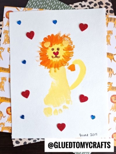 A HANDPRINT FATHER'S DAY lion craft made from a yellow painted footprint with an orange mane, black nose, and small eyes. Red glitter hearts and blue jewels decorate the white paper on lion-themed paper. Text: @GLUEDTOMYCRAFTS.