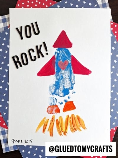 Children’s painting of a red and blue rocket ship with orange flames, a heart, and the words “YOU ROCK!” on white paper—perfect for a HANDPRINT FATHER'S DAY craft—placed on star-patterned backgrounds. ©GLUEDTOMYCRAFTS is at the bottom.