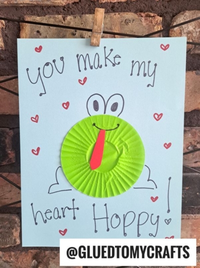 A handmade cupcake liner card features a green frog with a red tongue on blue paper. The card reads, "You make my heart HOPPY!" with small red hearts drawn around the text. @GLUEDTOMYCRAFTS is printed at the bottom—perfect for card crafts lovers!.