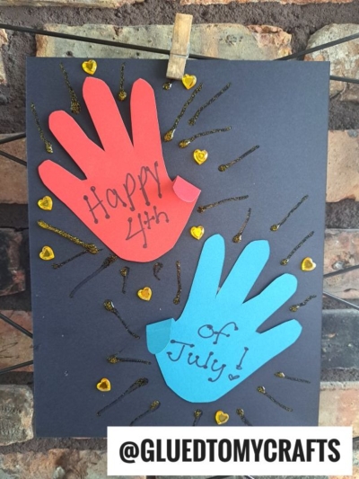 A craft with red and blue paper handprints on black paper, decorated with gold glitter and small yellow hearts—perfect for 4th of July keepsakes! The red hand says Happy 4th and the blue one says of July! @GLUEDTOMYCRAFTS is at the bottom.
