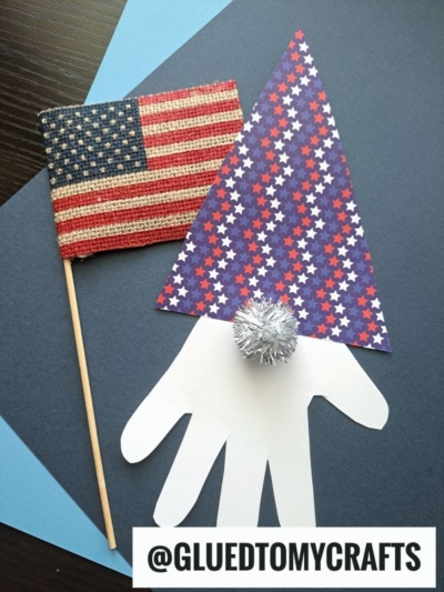 A patriotic craft featuring a handprint cutout with a blue star-covered triangle hat, a silver pom-pom nose, and a small American flag on a wooden stick—perfect for 4th of July keepsakes. The username @GLUEDTOMYCRAFTS is at the bottom.
