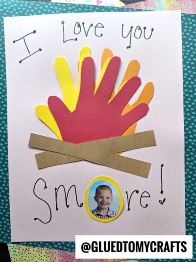 A handmade HANDPRINT FATHER'S DAY card features layered handprint cutouts in red, orange, and yellow, two brown paper strips crossed like logs, a child's photo, and decorated letters spelling "I Love You SMORE!.