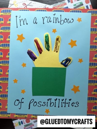 A handmade back to school keepsake displays a yellow handprint with each finger painted in different colors, emerging from a green rectangle on blue paper. Text reads "I'm a rainbow of possibilities," surrounded by orange stars.