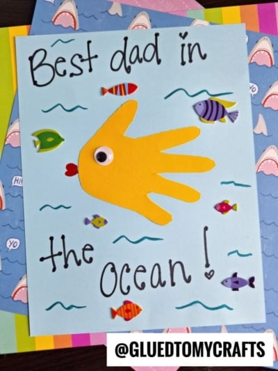 A handmade HANDPRINT FATHER'S DAY card with “Best dad in the ocean!” written on it. It features a yellow handprint fish with a googly eye and red lips, surrounded by colorful fish stickers.