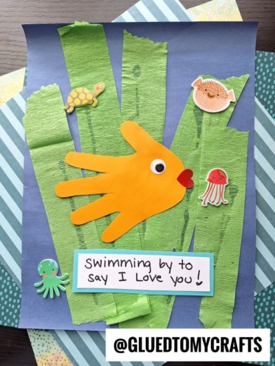 A HANDPRINT FATHER'S DAY fish made from an orange handprint is surrounded by seaweed and ocean animal stickers on blue paper. A note says “Swimming by to say I love you!” The image is labeled @gluedtomycrafts.