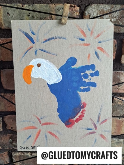 A handmade parrot painting created using a blue handprint, with orange, white, and red details—perfect for 4th of July keepsakes—is clipped to a line against a brick wall. @GLUEDTOMYCRAFTS is labeled at the bottom.