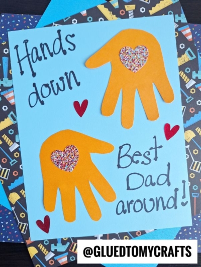 A handmade HANDPRINT FATHER'S DAY card with two orange paper handprints, each featuring a heart shape filled with colorful confetti. The card reads "Hands down, Best Dad around!" and is decorated with small red hearts.