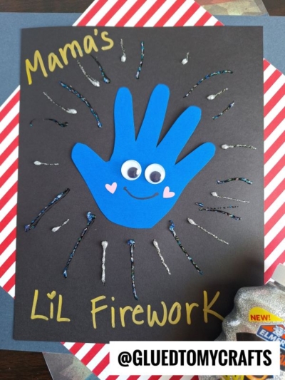 A handmade card features a blue paper handprint with googly eyes, heart cheeks, and a smile, surrounded by firework lines on a black background. Perfect for 4th of July keepsakes, it reads "Mamas LiL Firework.