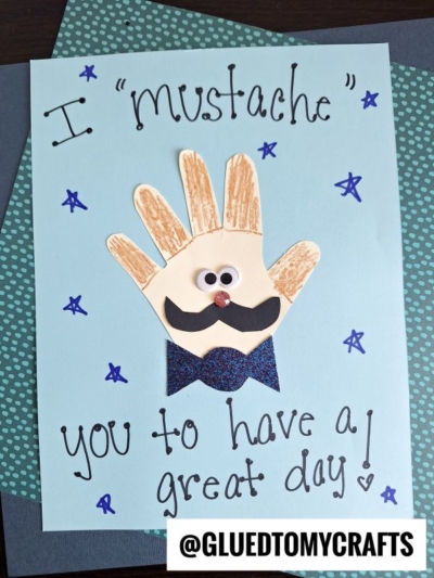 A handmade HANDPRINT FATHER'S DAY card featuring a cut-out handprint decorated with a mustache, googly eyes, and bow tie. Text reads “I ‘mustache’ you to have a great day!” with stars drawn around for extra fun.