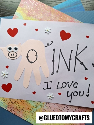 A handmade card features a pig made from a pink handprint cutout with googly eyes and a drawn snout, surrounded by red hearts and white flowers. Perfect for HANDPRINT FATHER'S DAY crafts, it reads “OINK I LOVE YOU!” and displays @GLUEDTOMYCRAFTS.