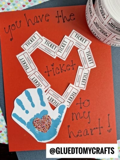 A handmade Valentine’s card on red paper features raffle tickets in a heart shape, handwritten You have the ticket to my heart!, and a blue handprint with a glittery heart—perfect inspiration for a HANDPRINT FATHER'S DAY card. A roll of tickets sits nearby.