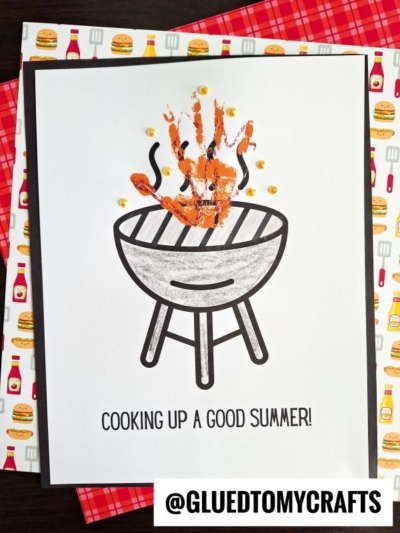 A paper craft features a grill topped with a painted handprint as flames and the phrase “COOKING UP A GOOD SUMMER!” below. Grilling-themed decorative paper in the background adds to this fun, summer-inspired project.