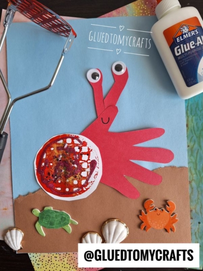 A kids’ craft project features a red handprint crab with googly eyes and a red circular mesh as a shell, on blue and brown paper, alongside sea animal stickers, shells, glue, and a kitchen masher.
