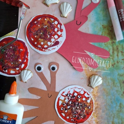 Two handprint crab crafts made from red and brown paper, decorated with googly eyes and glitter, are surrounded by seashells, glue, crayons, and crafting tools on a colorful background. The handle @GLUEDTOMYCRAFTS is shown.