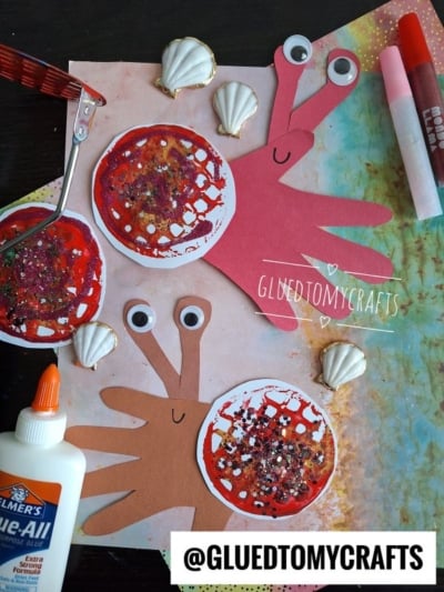 Two handprint crab crafts made from red and brown paper, decorated with googly eyes and glitter, are surrounded by seashells, glue, crayons, and crafting tools on a colorful background. The handle @GLUEDTOMYCRAFTS is shown.