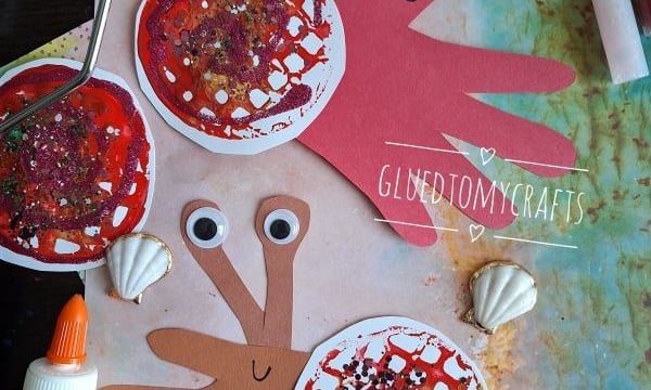 Two handprint crab crafts made from red and brown paper, decorated with googly eyes and glitter, are surrounded by seashells, glue, crayons, and crafting tools on a colorful background. The handle @GLUEDTOMYCRAFTS is shown.