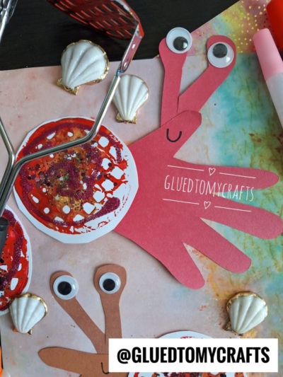 A handmade snail craft using a red paper handprint for the snail’s body and a painted round shell, surrounded by seashells, craft tools, and art supplies on a colorful background. @GLUETOMYCRAFTS is visible.