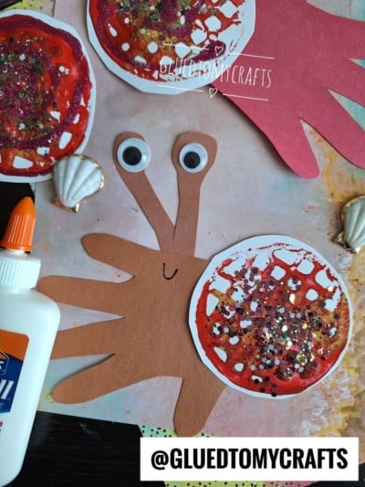 Handmade crab craft using cutouts of red and brown handprints for claws and body, googly eyes, a smiling face, decorated pincers with glitter, seashells, glue, and a @GLUEDTOMYCRAFTS label at the bottom.