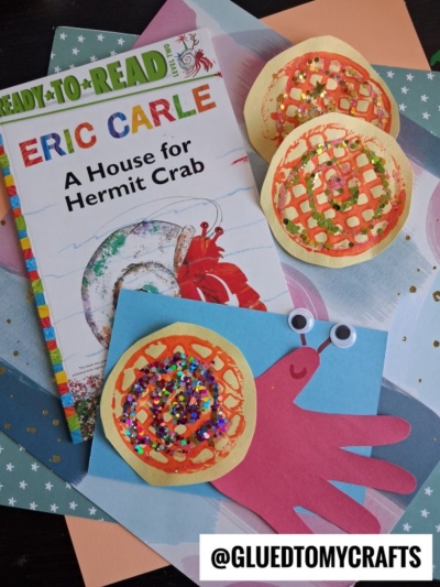 A children’s book, *A House for Hermit Crab* by Eric Carle, is surrounded by colorful crab crafts—some featuring a potato masher stamped hermit crab design—made from paper, glitter, and googly eyes, all arranged on decorated sheets. @GLUEDTOMYCRAFTS is in the corner.