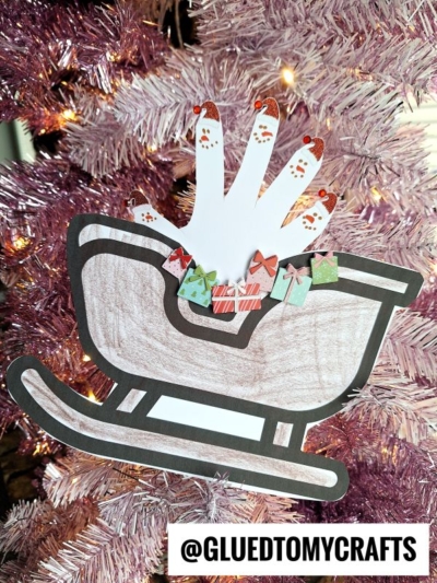 A handmade Christmas sleigh keepsake featuring Santa’s hand with painted fingernails, sitting in a colored sleigh filled with tiny paper gifts and attached to a pink Christmas tree. Text reads @GLUEDTOMYCRAFTS.
