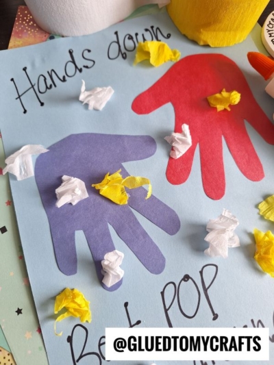 A handmade card with red and purple cut-out handprints on blue paper, decorated with crumpled yellow and white tissue paper. The text reads “Hands down, Best Pop.” The @GLUEDTOMYCRAFTS watermark is visible.