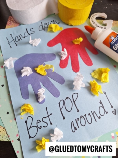A handmade card with cut-out handprints, glued tissue paper, and the message Hands down, Best POP around! sits on a table with a glue bottle, toilet paper roll, and craft supplies.