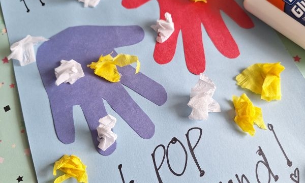 A handmade card with cut-out handprints, glued tissue paper, and the message Hands down, Best POP around! sits on a table with a glue bottle, toilet paper roll, and craft supplies.