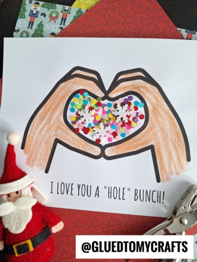 A drawing of two hands forming a heart shape holds colorful paper confetti. Below, the text reads "love you a hole bunch!" A small Santa figurine, holiday paper, and a hole puncher complete this festive scene.