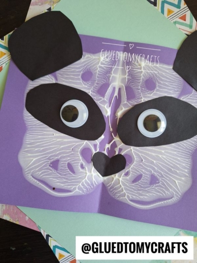 A handmade paint splat panda bear face card with large googly eyes, black paper ears, and a heart-shaped nose on a purple background. Decorative paper layers and @GLUEDTOMYCRAFTS watermark are also visible.