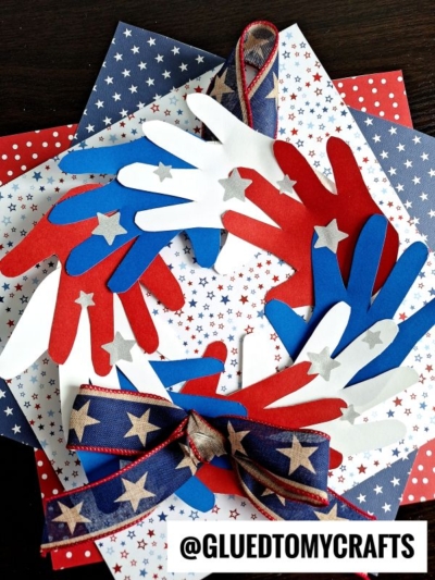 A patriotic wreath made of red, white, and blue paper hand cutouts with silver stars, layered on star-patterned paper, and decorated with a blue ribbon covered in stars and stripes—perfect for 4th of July keepsakes.