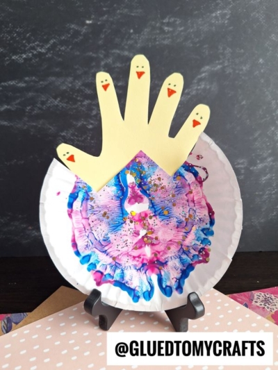 A colorful PAINT SPLAT CRAFT with blue, pink, and purple paint, topped with a yellow handprint cutout resembling a crown and four chicks. The Instagram handle @GLUEDTOMYCRAFTS is displayed at the bottom.