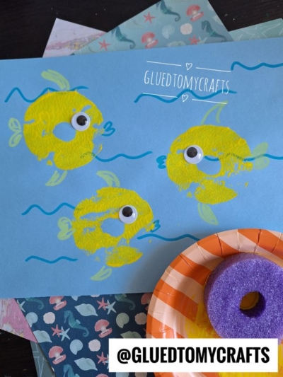 Blue paper with three yellow pool noodle stamped fish made from circles, each with a googly eye. Blue wavy lines represent water. A sliced purple pool noodle and a small orange tray are nearby. @GLUEDTOMYCRAFTS is written at the bottom.
